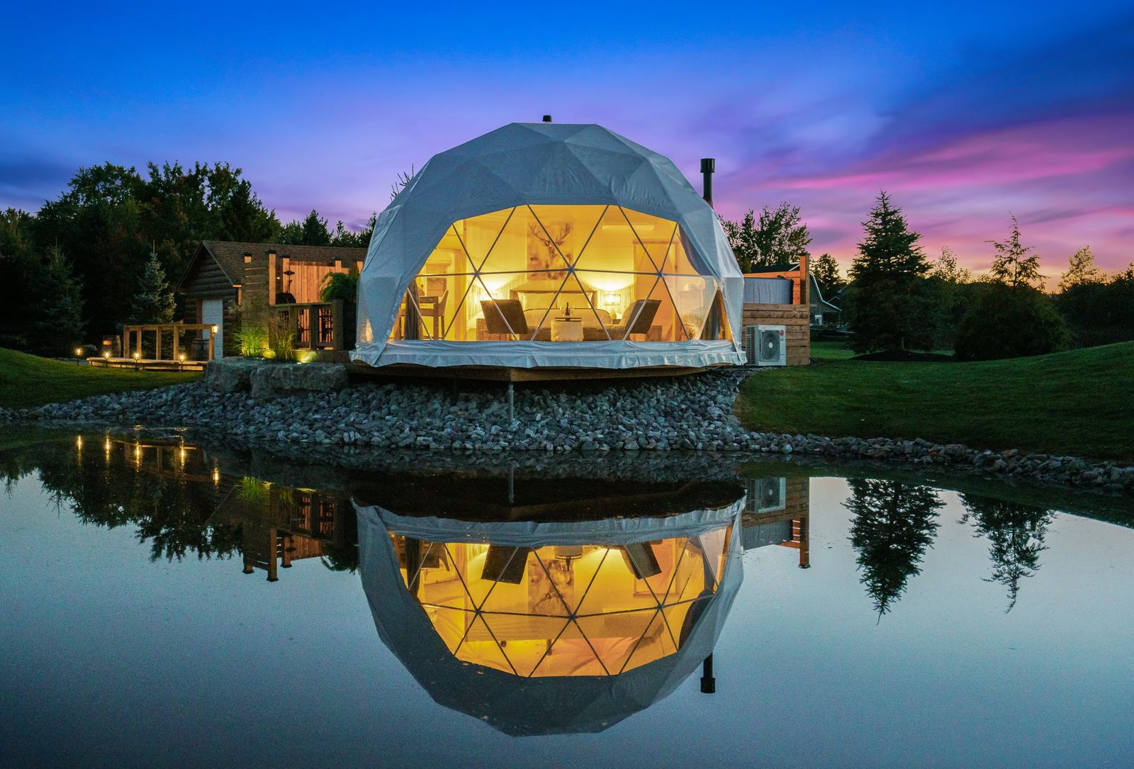 A lit geodesic dome beside still water at dusk, trees behind